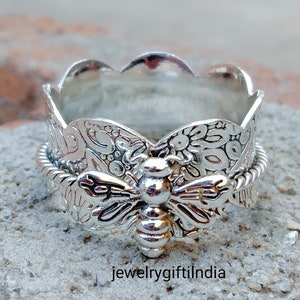 May include: A silver ring with a detailed bee design and a textured band. The bee is centered on the ring and has intricate wings and a textured body. The band is textured with a floral pattern.