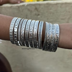 May include: A set of silver bangles with intricate designs. The bangles are stacked on a person's arm. The bangles have a variety of patterns, including geometric shapes, floral motifs, and abstract designs.