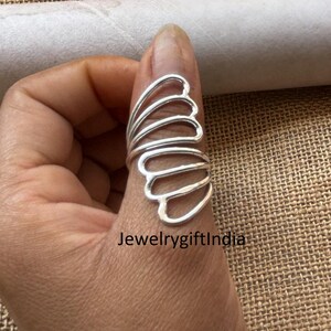 May include: A silver thumb ring with a delicate, layered design resembling a butterfly wing. The ring is made of thin, curved metal and has a minimalist aesthetic.