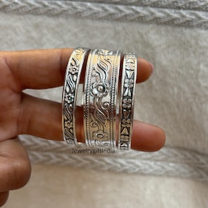 Könnte beinhalten: Set aus vier silbernen Armreifen mit filigranen Blumen- und geometrischen Mustern. Die Armreifen sind übereinander gestapelt und zeigen die verschiedenen Muster.