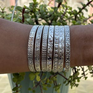May include: A set of silver bangles, each with a unique embossed design featuring floral, leaf, and geometric motifs. The bangles are displayed on a wrist, highlighting the detailed craftsmanship and diverse textures. The jewellery is set against a backdrop of green foliage.