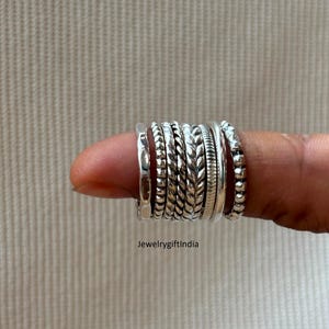 May include: A collection of silver rings with various textures, including braided, beaded, and smooth designs. The rings are stacked on a finger, showcasing the different styles. The image includes the text "JewelrygiftIndia".