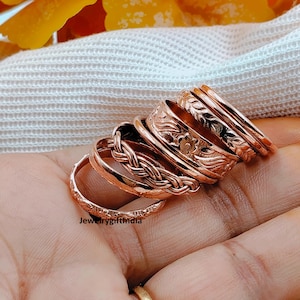 May include: A set of five copper-toned rings stacked together. The rings feature various textures including a braided pattern, a floral embossed design, and a simple hammered band. The text JewelrygiftIndia is visible on the bottom ring.
