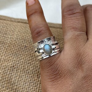 Könnte beinhalten: Ein silberner Ring mit einem blau-grauen ovalen Edelstein, der in der Mitte eingesetzt ist. Der Ring besteht aus mehreren Bändern mit einem filigranen Blumendesign.