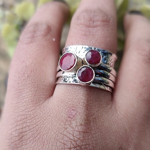 May include: A silver ring with three red gemstones set in a hammered band. The ring is stacked on top of three other silver bands.