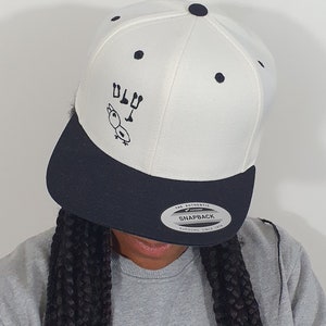 Pode incluir: Boné snapback branco e azul marinho com aba reta. O boné apresenta um desenho de pato de desenho animado e o texto "DLT" bordado em preto. É um boné Yupoong Snapback.