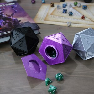 May include: Three 3D printed dice, one black, one purple, and one gray, with a purple lid and green dice on a wooden surface. The dice are shaped like a 20-sided die.