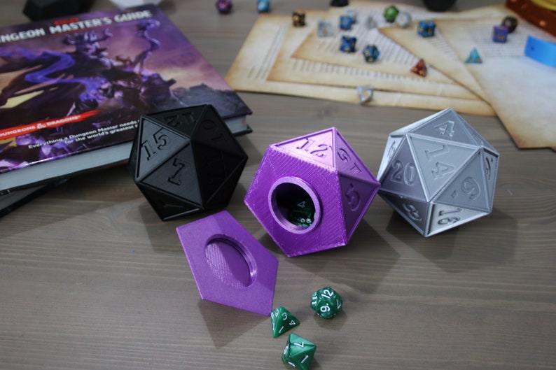 May include: Three 3D printed dice, one black, one purple, and one gray, with a purple lid and green dice on a wooden surface. The dice are shaped like a 20-sided die.