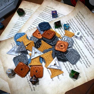Könnte beinhalten: Eine Sammlung verschiedener Spielmarken aus Kunststoff in Kupfer, Silber, Blau und Schwarz mit verschiedenen geprägten Symbolen. Die Spielmarken sind zusammen mit mehreren mehrseitigen Würfeln auf mehreren Blättern gealtertem Pergamentpapier verstreut.
