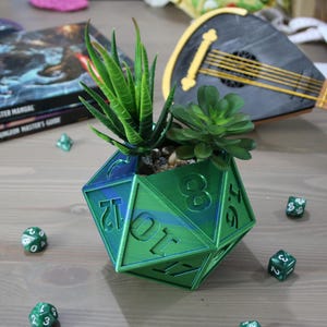 Op de afbeelding: Een groene, blauwe en zilveren, iriserende 20-zijdige dobbelsteenvormige plantenpot met twee kleine vetplanten erin. De pot staat op een houten oppervlak met verschillende groene dobbelstenen eromheen.