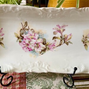 May include: A rectangular white porcelain tray with scalloped, embossed edges. The center features a painted floral motif with pink and white blossoms and green leaves, flanked by two smaller matching floral sprigs on either side of the tray.