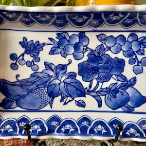 May include: A rectangular ceramic tray with a scalloped edge, featuring a white base and a blue hand-painted botanical design. The central illustration depicts clusters of berries, leaves, and flowers, while the border displays a repeating pattern.