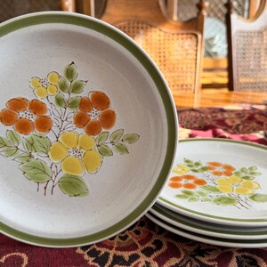 May include: A set of ceramic plates with a floral design. The plates have a speckled white background, a green rim, and a bouquet of orange and yellow flowers with green leaves in the center. Several plates are stacked.