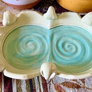 May include: A dual-well ceramic soap dish featuring a glossy cream-colored rim and six small decorative protrusions along the edge. The two interior basins are finished in a vibrant turquoise glaze with a distinct hand-carved spiral pattern.
