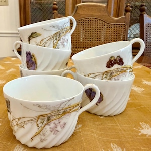 May include: A set of four white porcelain teacups featuring a delicate swirl texture on the exterior. Each cup is decorated with a painted design of brown and purple butterflies resting on thin, golden-brown branches with small floral accents.