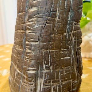 May include: A brown, textured ceramic vase with a rough, carved surface. The vase has a cylindrical shape with a slightly irregular top. The surface is marked with deep, linear grooves and a natural, earthy aesthetic. The vase is likely handmade.