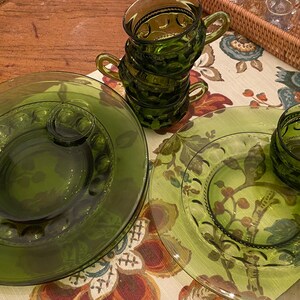 Puede incluir: Juego de cuatro tazas de vidrio verde y dos platillos a juego. Las tazas tienen un dise&ntilde;o en relieve y est&aacute;n apiladas una encima de la otra. Los platillos tienen un dise&ntilde;o similar y est&aacute;n colocados sobre un mantel floral.