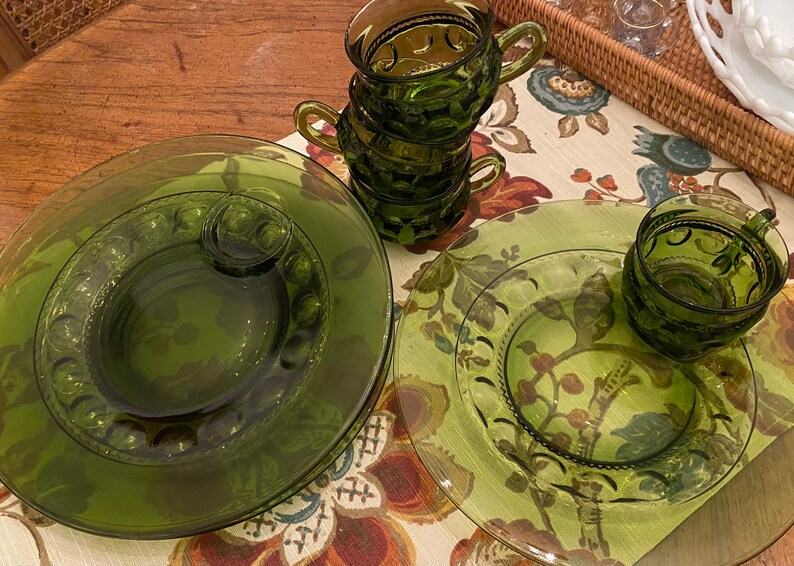 Puede incluir: Juego de cuatro tazas de vidrio verde y dos platillos a juego. Las tazas tienen un dise&ntilde;o en relieve y est&aacute;n apiladas una encima de la otra. Los platillos tienen un dise&ntilde;o similar y est&aacute;n colocados sobre un mantel floral.