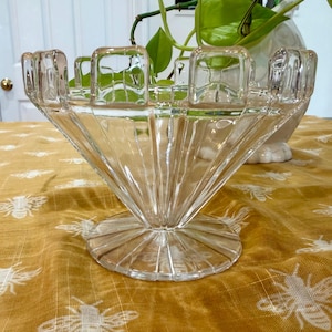 May include: A clear glass decorative bowl with a fan-shaped design. The bowl has a round base and a series of square-shaped holders around the top rim. The bowl is sitting on a yellow tablecloth with a bee pattern.
