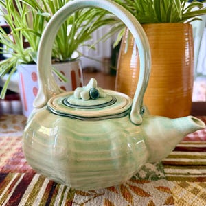 May include: A light green ceramic teapot with a glossy finish and a high arched handle. The lid features a small sculpted floral knob with a dark teal center. The body has a ribbed texture and a short spout, showcasing a hand-crafted appearance.