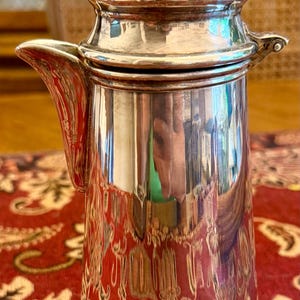 May include: A polished silver-toned metal coffee pot featuring a cylindrical body, a curved spout, and a domed lid topped with a small, rounded finial. The reflective surface of the metal shows distorted surroundings and a smooth, metallic finish.