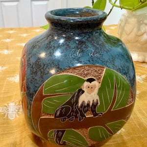 May include: Blue ceramic vase with a textured finish and a detailed illustration of a monkey on a tree branch. The vase is inscribed with "Costa Rica". The vase has a wide base and a narrow neck.