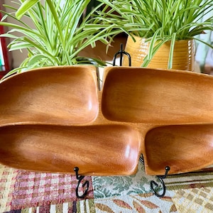 May include: A light brown wooden serving tray shaped like a leaf with four distinct, smooth, concave compartments. The tray features a natural wood grain finish and a small handle on the right side, supported by a black metal display stand.