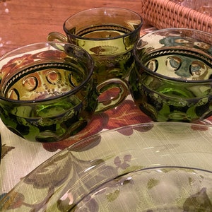 Puede incluir: Tres tazas de vidrio verde con un dise&ntilde;o texturizado y un borde festoneado. Las tazas tienen un asa y est&aacute;n sobre una superficie con estampado floral.