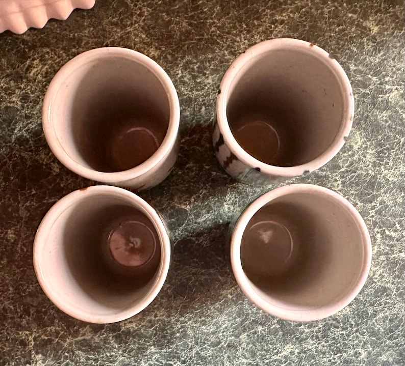 Puede incluir: Cuatro tazas de cer&aacute;mica blanca con esmalte moteado marr&oacute;n y negro. Las tazas est&aacute;n vac&iacute;as y est&aacute;n sobre una superficie marr&oacute;n.