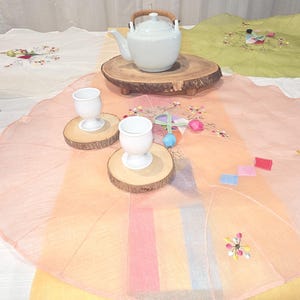 May include: A white ceramic teapot with a wooden handle sits on a wooden slab. Two white egg cups rest on wooden coasters, with a colorful, embroidered tablecloth in the foreground. The tablecloth has a floral design.