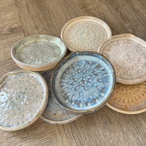 Könnte beinhalten: Eine Sammlung handgefertigter Keramik-Reibeplatten in verschiedenen Farben, darunter Blau, Beige und Grün. Jede Platte hat eine strukturierte Oberfläche mit einem einzigartigen Muster aus kleinen, erhabenen Noppen. Die Platten sind auf einer Holzoberfläche angeordnet.