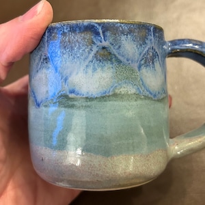 May include: A ceramic mug with a rounded body and a large handle. The mug has a colour gradient, going from dark blue at the top to light blue and beige at the base. The mug is held by a hand.