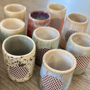 May include: A selection of handmade ceramic shot glasses in various colours, including beige, green, and red. Each glass features a unique speckled pattern and a honeycomb-like design. The glasses are cylindrical and appear to be glazed.