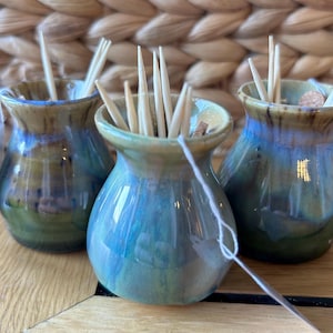May include: Three small, handmade ceramic vases with a blue, green, and brown glaze. Each vase is filled with wooden toothpicks. The vases have a unique, organic shape and are displayed on a wooden surface.