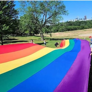 Puede incluir: Una gran bandera arcoíris del orgullo está siendo sostenida por un grupo de personas en un parque. La bandera está hecha de una tela colorida con los colores del arcoíris. Las personas están sonriendo y divirtiéndose.
