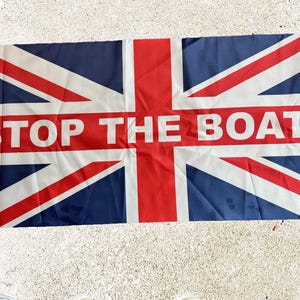 Könnte beinhalten: Eine rechteckige Flagge mit dem Design des Union Jack, mit einem roten und weißen Banner in der Mitte. Das Banner trägt die Aufschrift "STOP THE BOATS" in weißen Großbuchstaben. Die Flagge ist hauptsächlich blau, rot und weiß.