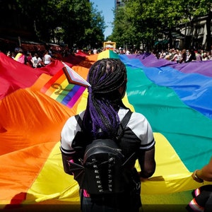 May include: A large rainbow pride flag is held up by a group of people during a parade. The flag is made of six different colors: red, orange, yellow, green, blue, and purple. The flag is held up high in the air, and the people are smiling and waving.
