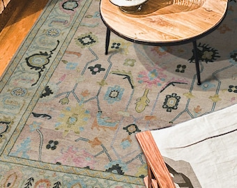 May include: An overhead shot of a living room featuring a patterned area rug with a light pink and blue color scheme. A round wooden coffee table sits on the rug, and two wooden chairs are placed around the table. A leather sofa is visible on the right.