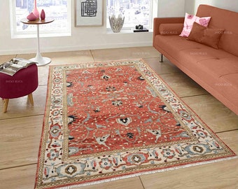 May include: A rectangular, red area rug with an intricate floral pattern and a cream-colored border. The rug features blue and black accents within the design. The rug is placed on a light-colored wooden floor, adding warmth to the room.