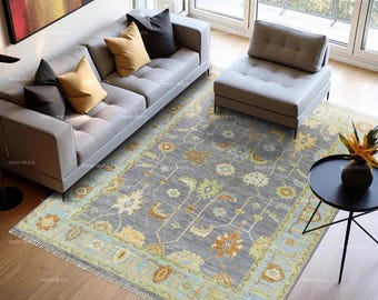 May include: A gray area rug with a floral pattern, featuring shades of yellow, blue, and green. The rug is placed in a living room setting with a gray sofa, pillows, and a small black table. The rug has a fringe.