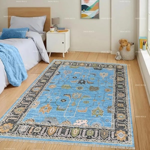 Pode incluir: Um tapete retangular azul claro com um padrão floral e uma borda escura. O tapete apresenta um design central com várias formas florais e geométricas em tons de laranja, verde e cinza. O tapete é colocado sobre um chão de madeira.
