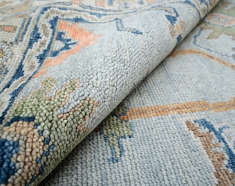 May include: Close-up of a rolled-up area rug with a detailed pattern. The rug features a light blue background with accents of navy blue, peach, and green. The design includes geometric and floral elements.