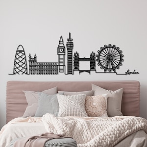 K&ouml;nnte beinhalten: Schwarze Metallsilhouette der Londoner Skyline, einschlie&szlig;lich London Eye, Big Ben und Shard. Die Silhouette ist an einer wei&szlig;en Wand &uuml;ber einem Bett mit einem rosafarbenen Kopfteil und wei&szlig;en Bettw&auml;sche montiert.
