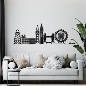 K&ouml;nnte beinhalten: Schwarze und wei&szlig;e Silhouette der Londoner Skyline, einschlie&szlig;lich Big Ben, London Eye und Shard. Die Skyline ist an einer wei&szlig;en Wand &uuml;ber einem grauen Sofa mit Kissen montiert. Das Wort "London" steht in Schwarz unter der Skyline.