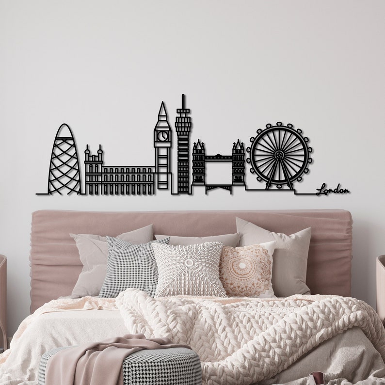 K&ouml;nnte beinhalten: Schwarze Metallsilhouette der Londoner Skyline, einschlie&szlig;lich London Eye, Big Ben und Shard. Die Silhouette ist an einer wei&szlig;en Wand &uuml;ber einem Bett mit einem rosafarbenen Kopfteil und wei&szlig;en Bettw&auml;sche montiert.