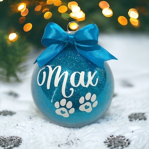 May include: A teal blue glitter Christmas bauble with a white bow and the name "Max" written on it. Two white paw prints with a heart in each are also on the bauble.