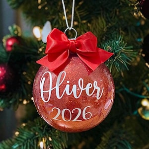 May include: A round, red glitter Christmas bauble featuring a white script name Oliver and the year 2026 printed in white. The bauble is topped with a bright red satin ribbon bow and hangs from a silver string loop for display purposes.