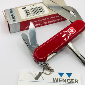 Pode incluir: Um canivete suíço Wenger vermelho com uma cruz branca no cabo. O canivete possui uma variedade de ferramentas, incluindo uma lâmina, um abridor de latas, um abridor de garrafas, uma chave de fenda e um saca-rolhas. O canivete é embalado em uma caixa vermelha com o logotipo da Wenger e o texto "Le Véritable Couteau" em francês.