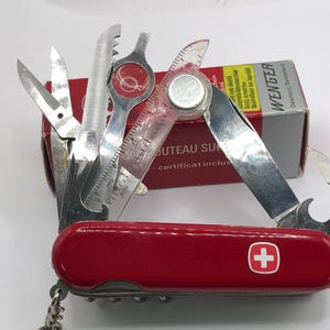 Op de afbeelding: Een rood Zwitsers zakmes met meerdere uitgevouwen gereedschappen, waaronder een schaar, een zaag en een liniaal. Het mes heeft een zilveren lemmet en een rode handgreep met een wit kruis embleem. Een kleine doos met het woord "Wenger" staat op de achtergrond.