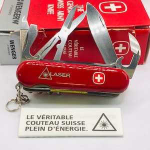 Può includere: Un coltellino svizzero Wenger rosso con diversi utensili in acciaio inossidabile aperti, tra cui una lama, forbici e un apriscatole. Il manico presenta un logo con croce bianca e la scritta LASER. È presentato con la scatola rossa originale.